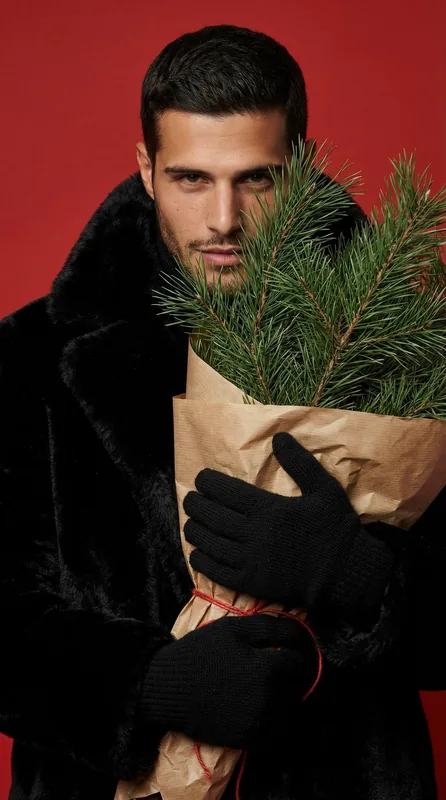 Editorial winter portrait shot against a bold red studio backdrop. A man in a textured black faux-fur coat stands center
